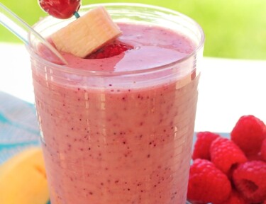 Banana berry smoothie in a glass with a cherry and banana slice on a toothpick and a bowl of raspberries in the background
