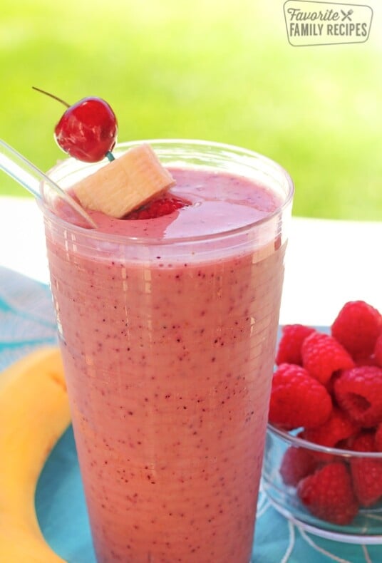 Banana berry smoothie in a glass with a cherry and banana slice on a toothpick and a bowl of raspberries in the background