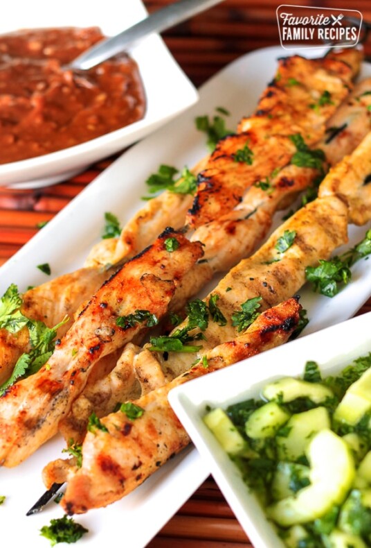 Chicken Satay on a white tray with Peanut Dipping Sauce and Cool Cucumber Salad on the side
