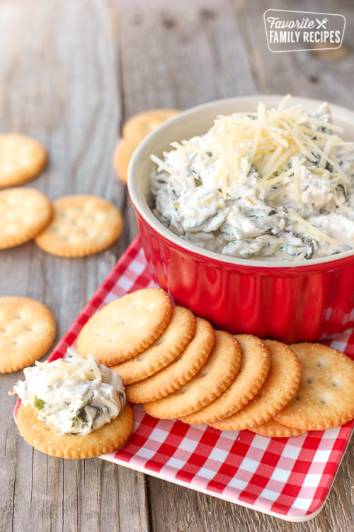 A hand holding Easy Spinach Dip on a Ritz cracker with a bowl of spinach dip in a red bowl in the background
