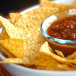 Homemade Salsa in a glass bowl surrounded by tortilla chips in a bigger white bowl.