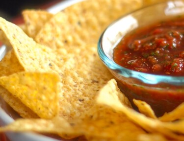 Homemade Salsa in a glass bowl surrounded by tortilla chips in a bigger white bowl.