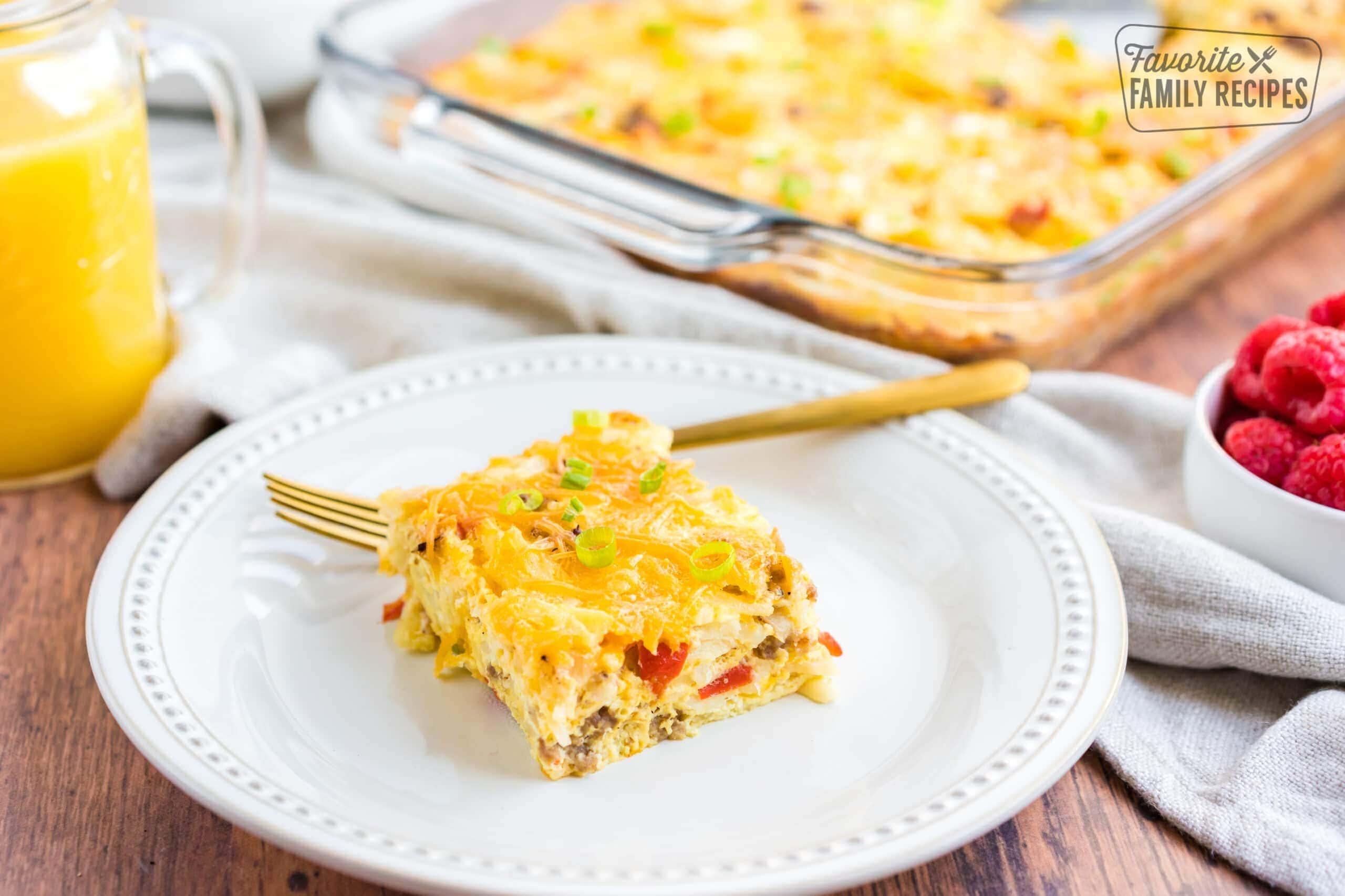 A square of breakfast casserole with egg, sausage, hash browns, tomato and green onion on a white plate.