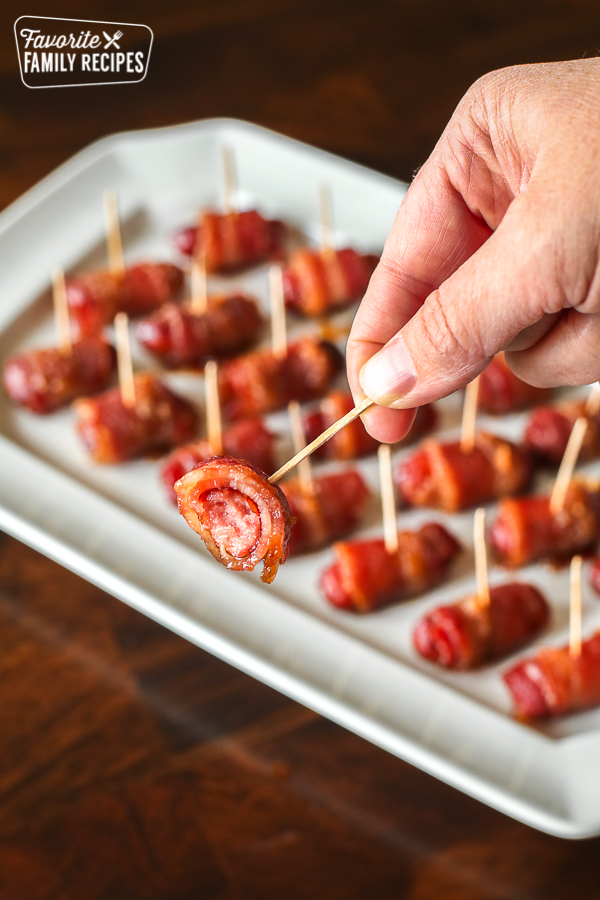 A hand holding a portion of a bacon wrapped lil smokies on a toothpick with a tray of Lil Smokies in the background
