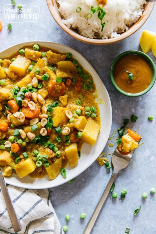 Coconut Curry Chicken with Peanuts, Potatoes, Carrots, and Peas in a white bowl with a bowl of rice and peanut sauce on the side