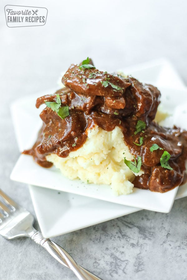 Crockpot steak and gravy served over mashed potatoes on a square, white plate.