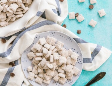 Muddy Buddies on a plate surrounded by chocolate chips and a spoonful of peanut butter.