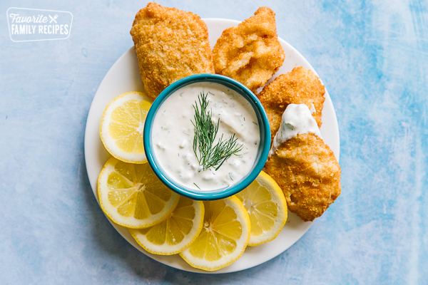 Tartar sauce with a sprig of dill in the middle on a plate with lemon wedges and fried fish