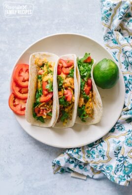 Three chicken tacos on a plate with sliced tomatoes and a lime on the side