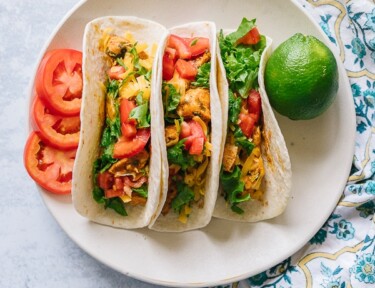 Three chicken tacos on a plate with sliced tomatoes and a lime on the side