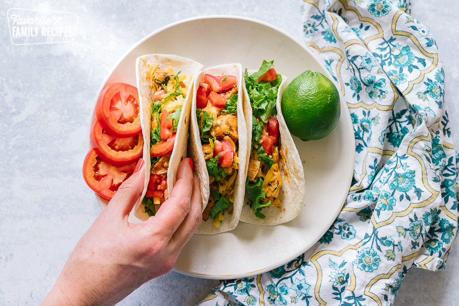 A hand reaching for one of three chicken tacos on a plate