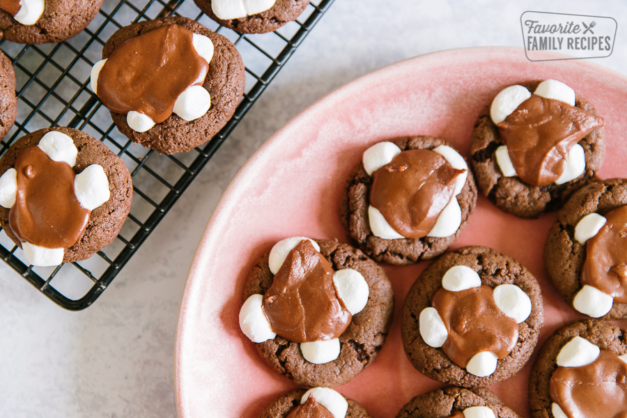 A pink plate filled with chocolate marshmallow cookies and a cooling rack with more cookies on the side