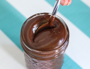 A spoon being lowered into a glass jar filled with hot fudge sauce