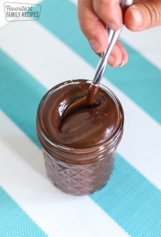 A spoon being lowered into a glass jar filled with hot fudge sauce