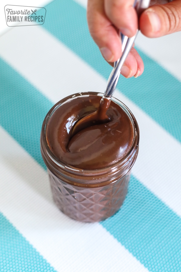 A spoon being lowered into a glass jar filled with hot fudge sauce.