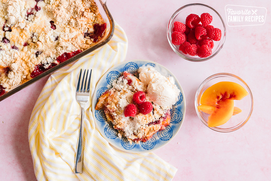 A serving of Raspberry Peach Cobbler on a blue plate with a casserole dish of cobbler on the side
