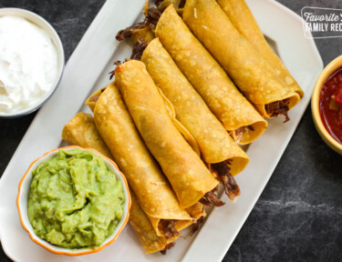 A platter of shredded beef taquitos with guacamole, sour cream, and salsa on the side