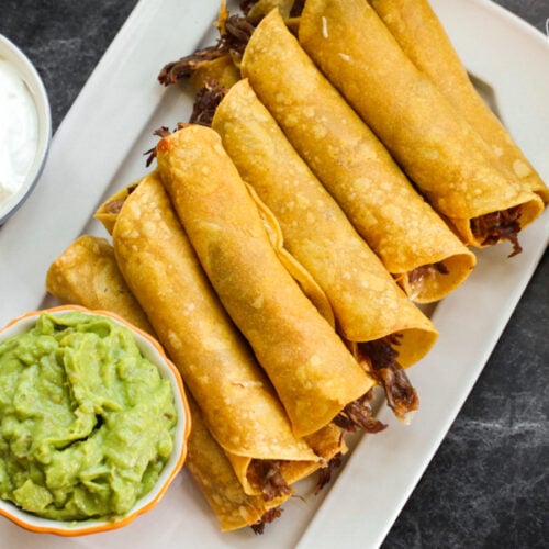 A platter of shredded beef taquitos with guacamole, sour cream, and salsa on the side
