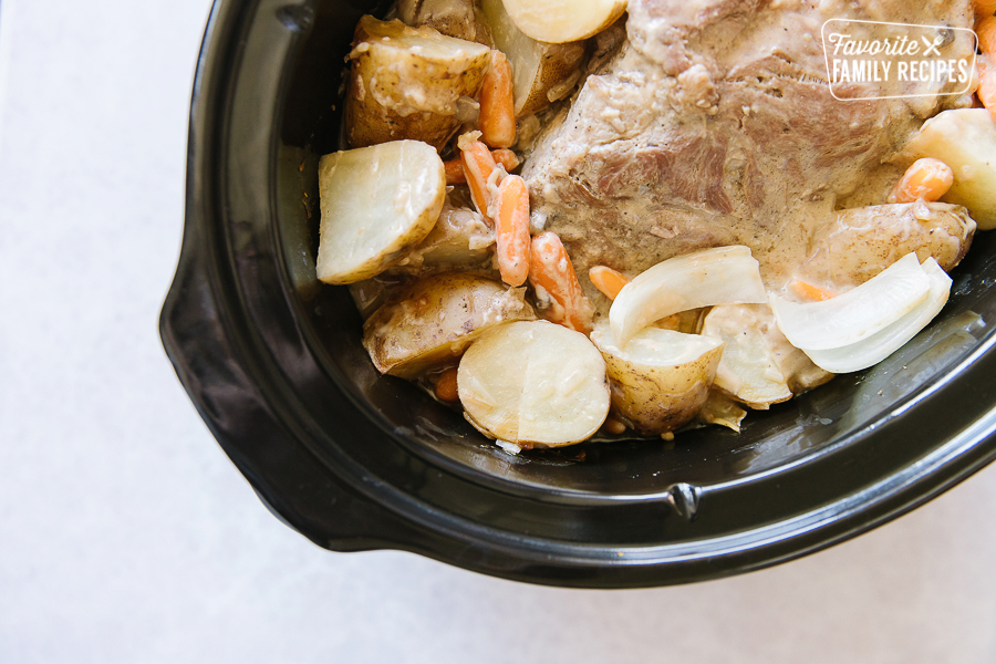 Pork Roast in a black crock pot surrounded by potatoes, carrots, onions.