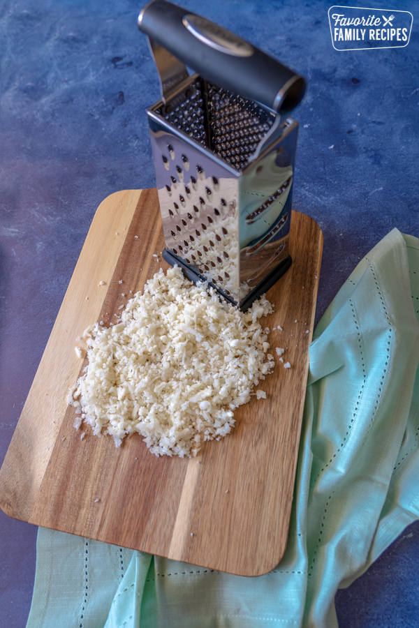 Riced Cauliflower on a cutting board next to a box grater. 