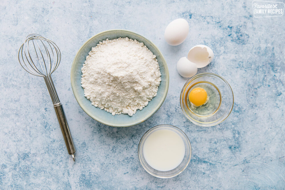 A whisk, A bowl of basic cake mix, an egg, and a bowl of milk on a blue background.