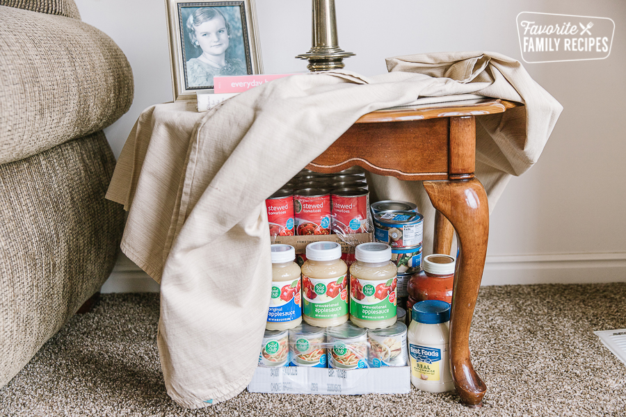 Cans of food stored under an end table with a tablecloth over it.