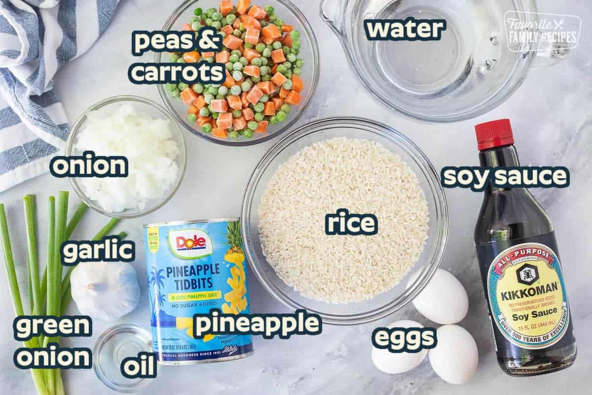 Ingredients to make pineapple fried rice including rice, soy sauce, eggs, water, peas and carrots, onion, pineapple tidbits, green onion, garlic and oil.