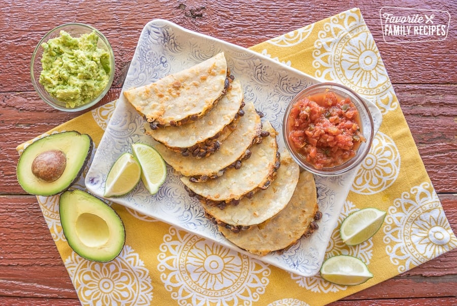 Black bean tacos stacked on a plate with a bowl of guacamole and a bowl of salsa