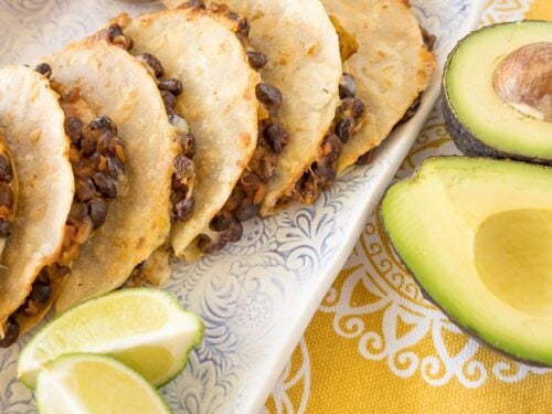 Black bean tacos stacked up on a plate with limes, avocados, and a bowl of salsa.
