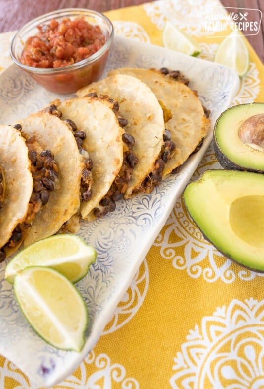 Black bean tacos stacked up on a plate with limes, avocados, and a bowl of salsa.