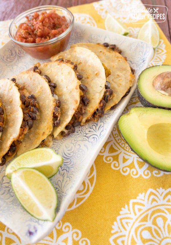 Black bean tacos stacked up on a plate with limes, avocados, and a bowl of salsa.
