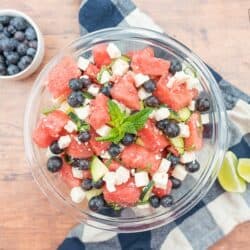 Watermelon feta salad in a glass bowl with limes and a bowl of blueberries