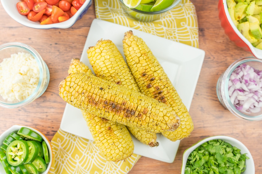 grilled corn on a plate with all the salad ingredients in little bowls around it
