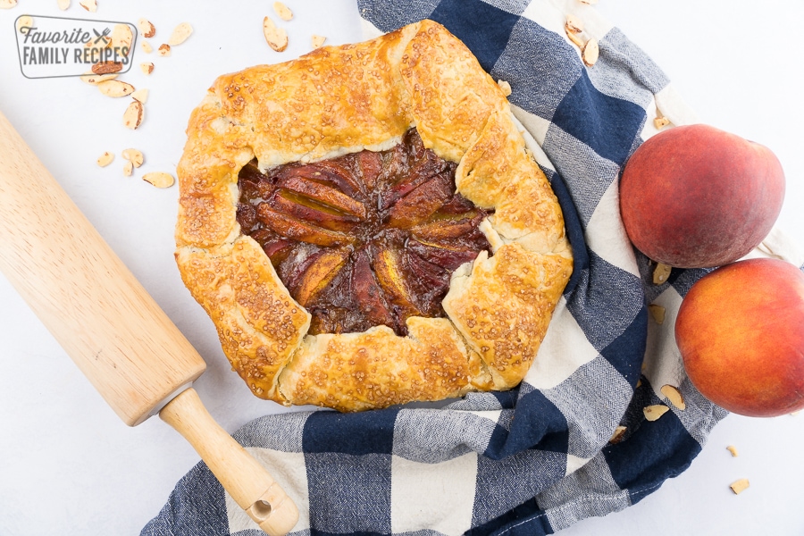 a peach galette on a blue checked napkin with a rolling pin and peaches