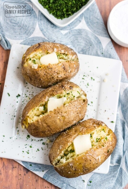 Three baked potatoes with butter and herbs and a bowl of herbs