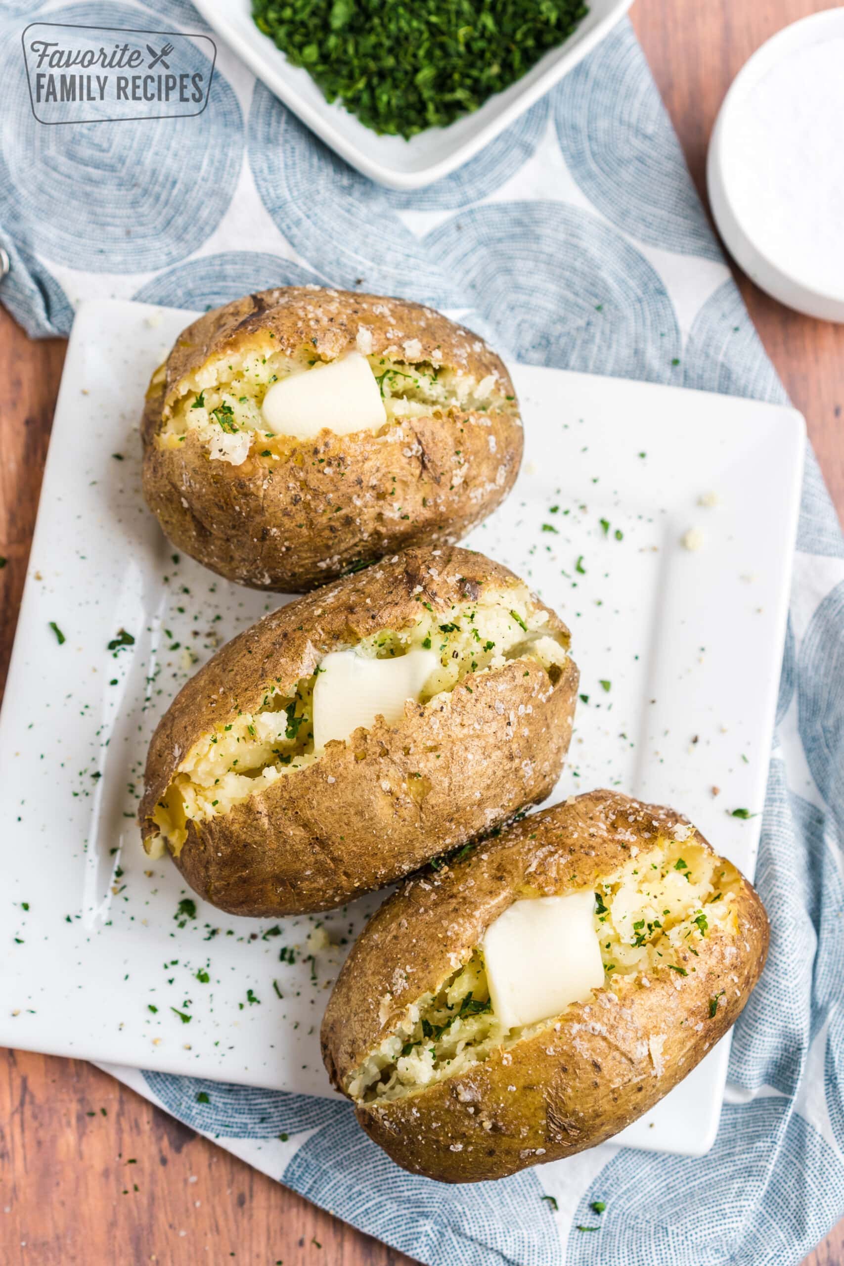 Three baked potatoes with butter and herbs and a bowl of herbs