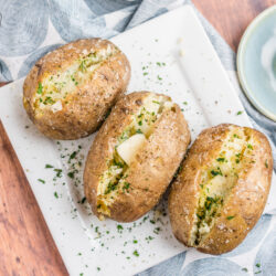 Three baked potatoes on a plate topped with butter and herbs