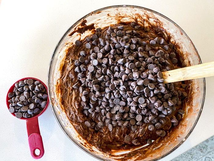 Chocolate zucchini dough in a glass bowl mixed in with chocolate chips with a wooden spoon.