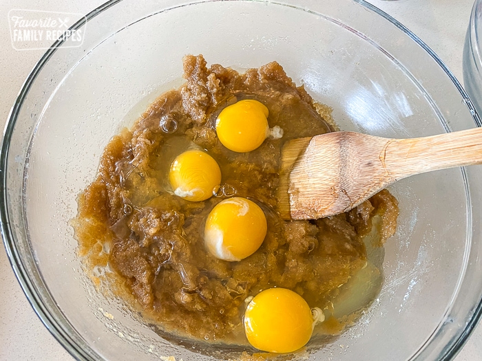 Eggs, brown sugar, and oil in a glass mixing bowl with a wooden spoon.