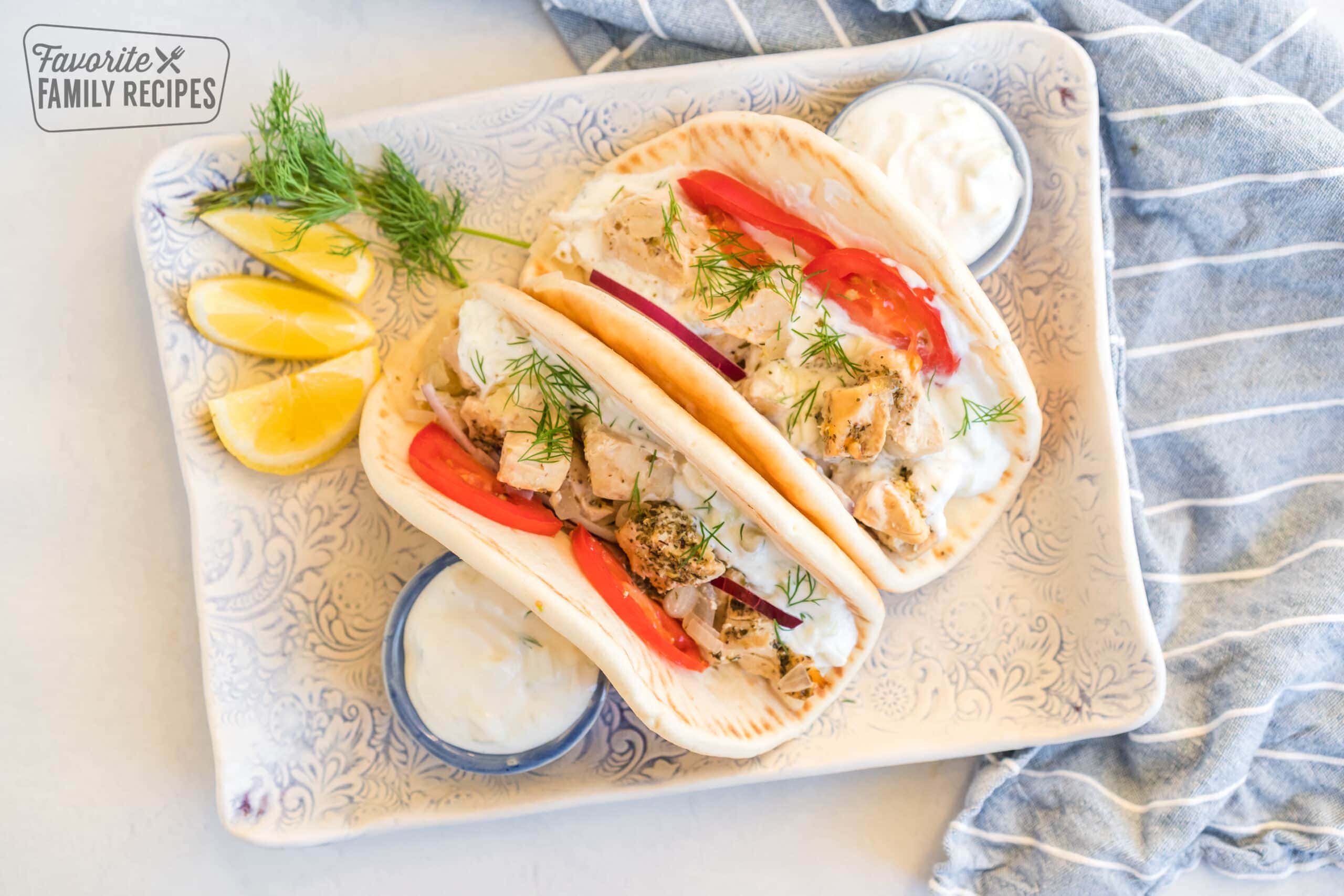 Two chicken gyros on a platter with bowls of tzatziki sauce, lemons, and dill 