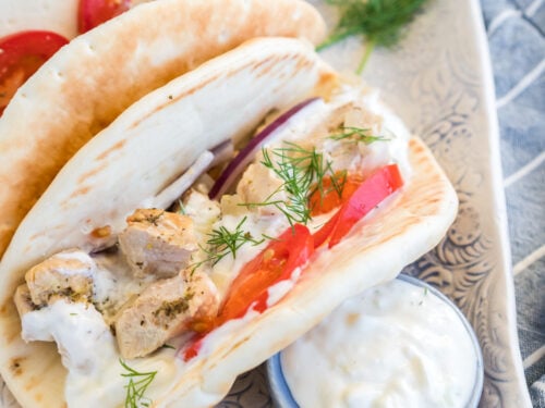 Two chicken gyros on a plate with a dish of tzatziki sauce, lemons, and dill