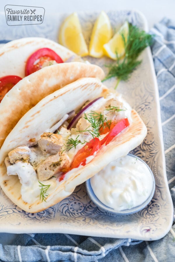 Two chicken gyros on a plate with a dish of tzatziki sauce, lemons, and dill