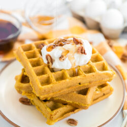 A stack of pumpkin waffles on a plate with whipped cream, pecans, and syrup