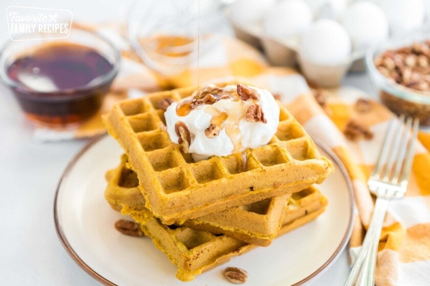 A stack of pumpkin waffles on a plate with whipped cream, pecans, and syrup