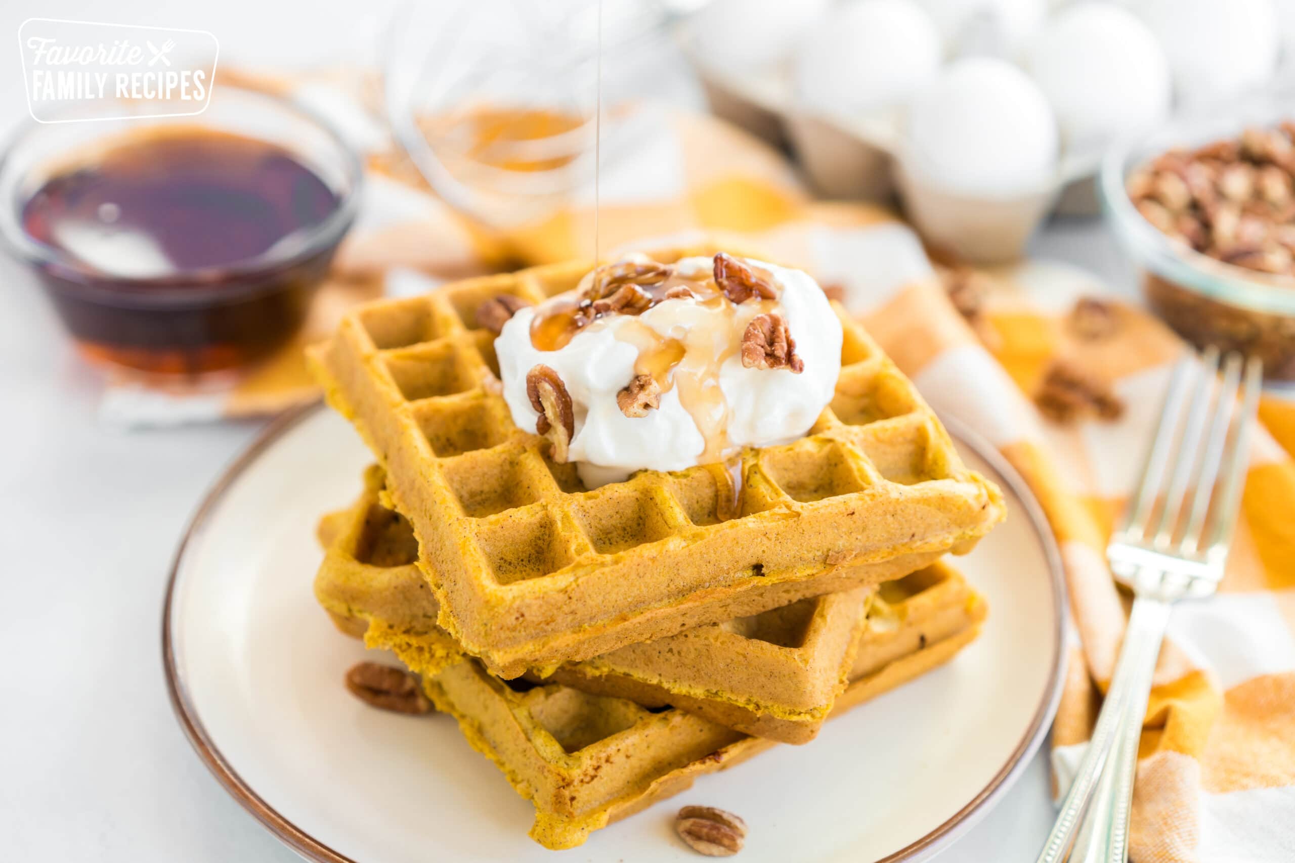 A stack of pumpkin waffles on a plate with whipped cream, pecans, and syrup.