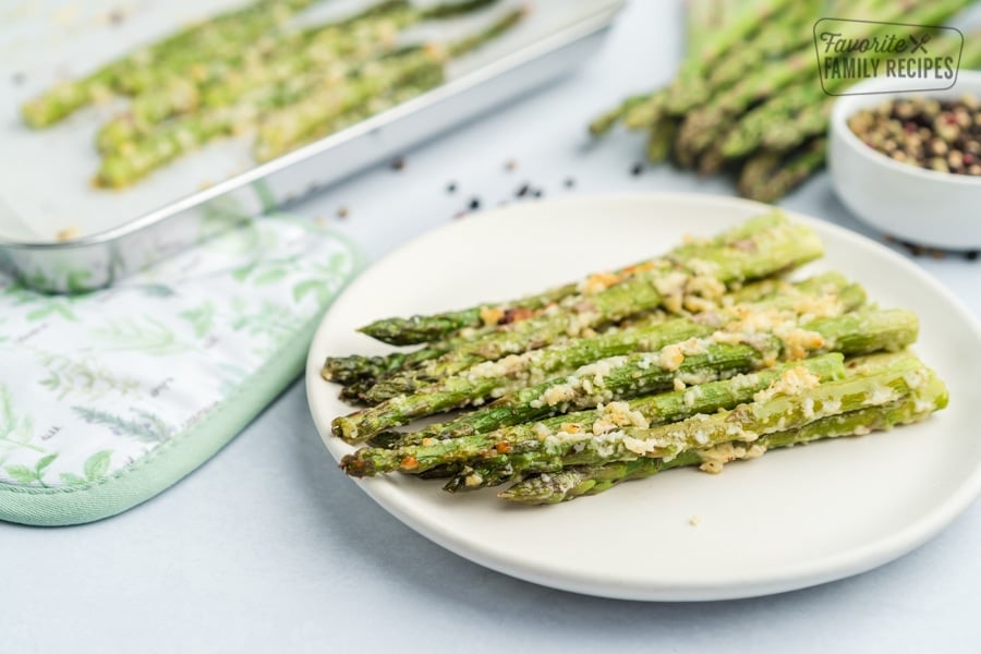 Oven Roasted Asparagus is a flavorful side dish for any meal. Topped with olive oil, garlic and cheese, they come out perfectly tender and crisp every time! Roasted asparagus on a white plate topped with garlic and cheese with a roasting pan in the background.