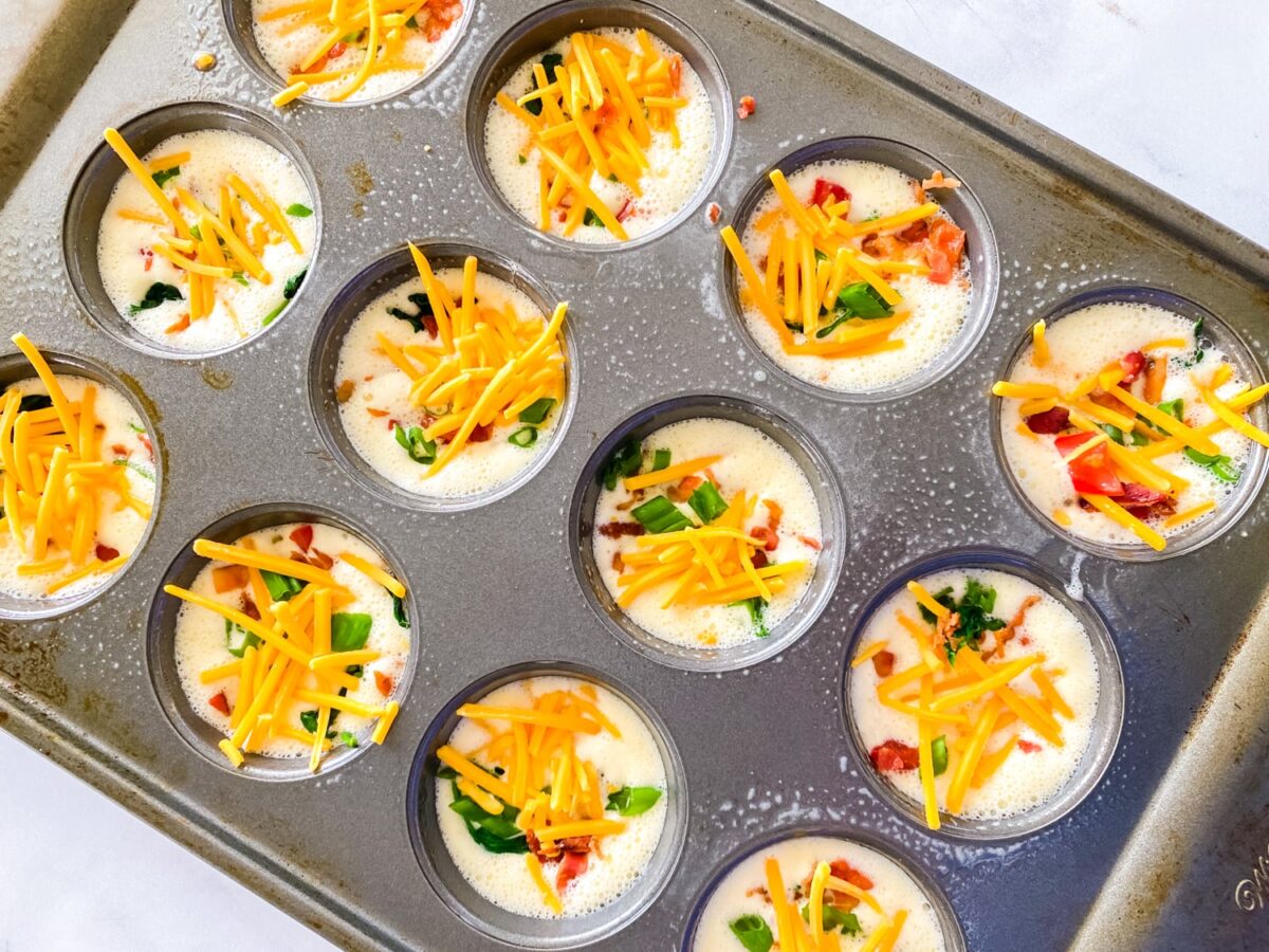 Eggs in a muffin tin topped with cheese, bacon, and green onion ready to be baked