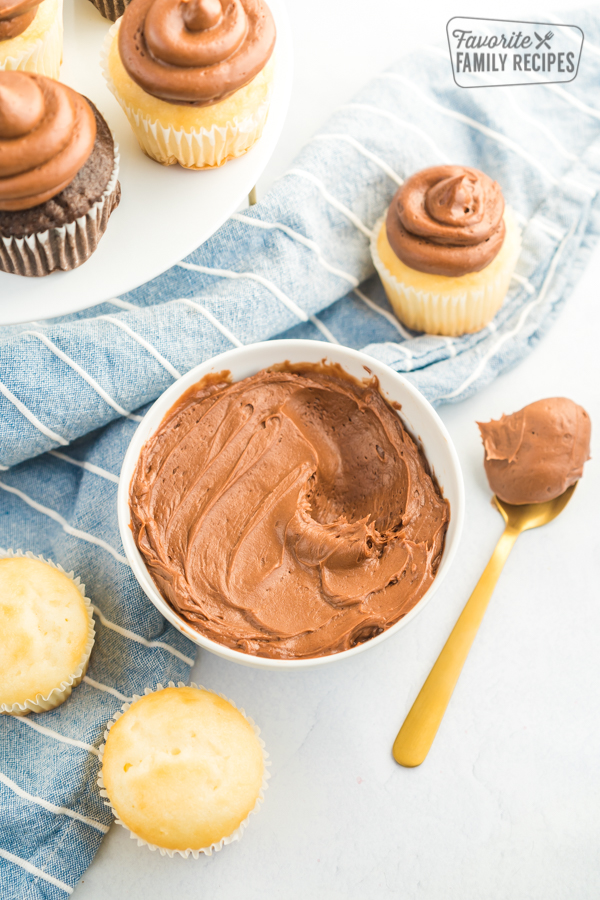 A bowl of chocolate buttercream frosting with a tray of cupcakes next to it