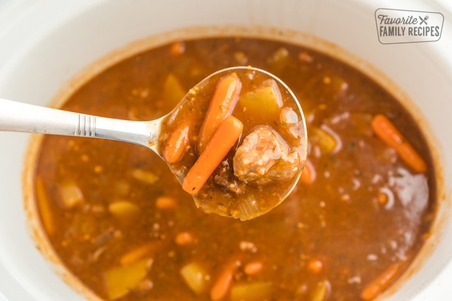 A scoop of beef stew in a ladle in front of a crock pot full of stew