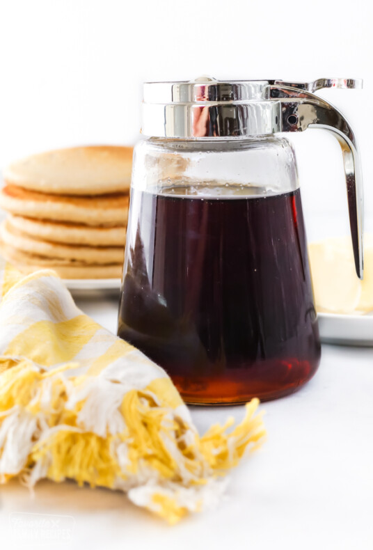 A jar of homemade maple syrup in front of a stack of pancakes and softened butter